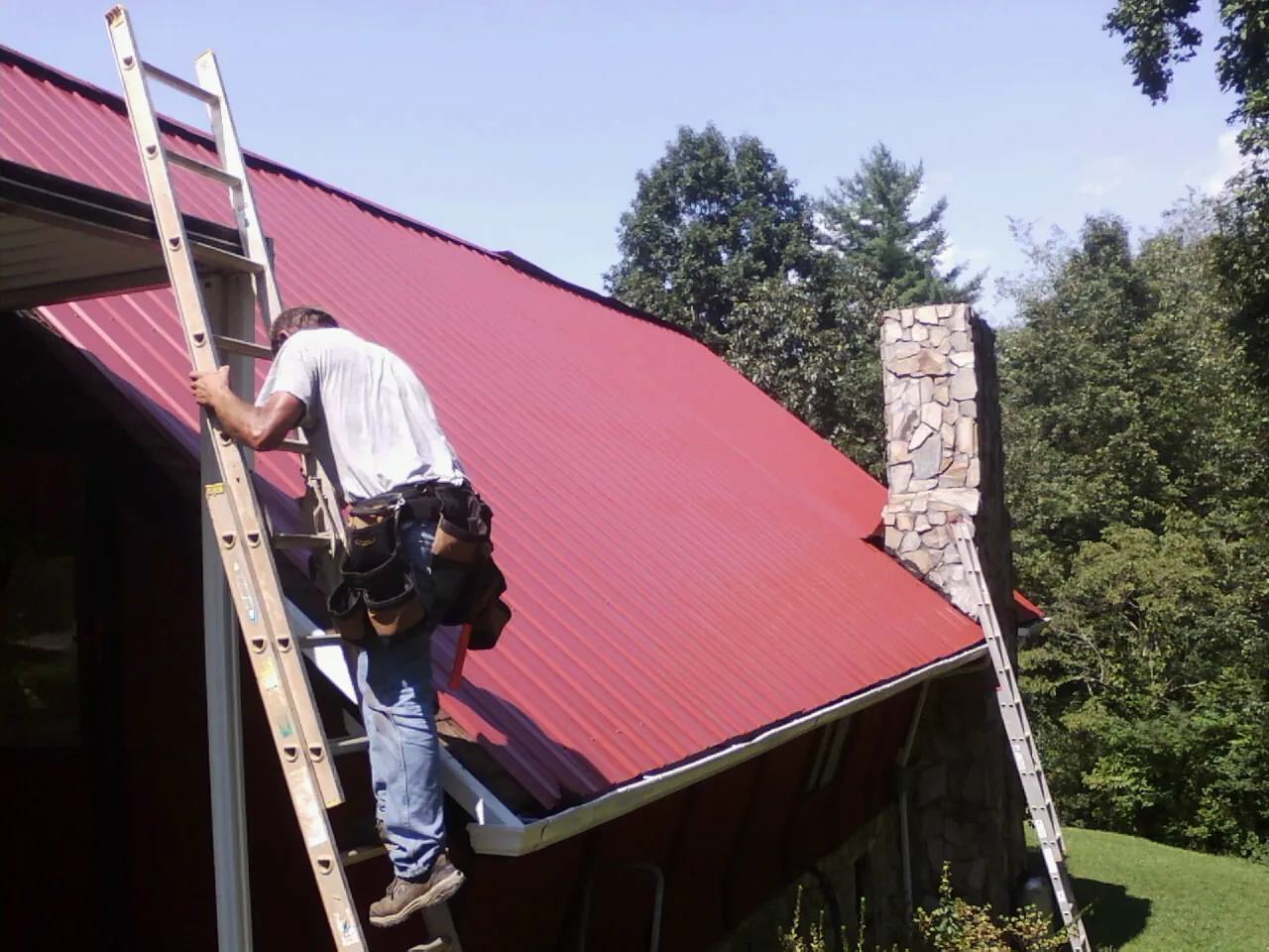 Roofer on a ladder beside a red metal roof and stone chimney — Soffit & Fascia Repair by Roofing Brothers in Ford Cliff